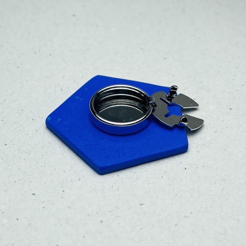 May include: A silver-colored metal bottle stopper with a circular design, attached to a blue, diamond-shaped base. The stopper has a unique, stylized shape. The background is a light gray surface.