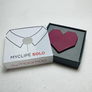 May include: A burgundy heart-shaped accessory rests in a gray box, next to a white box with a shirt collar graphic and the text "MYCLIPE SOLO by moomoo". The heart-shaped item is a fashion accessory.
