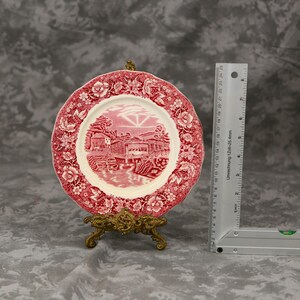 Pode incluir: Um prato decorativo com uma borda floral vermelha e branca e uma cena de paisagem central. O prato é exibido em um suporte dourado. Uma régua mede aproximadamente 23 cm.