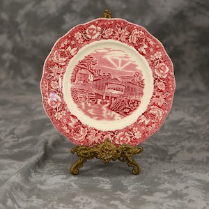 Peut inclure: Une assiette décorative avec une bordure florale rouge et une scène centrale d'un bâtiment et de l'eau. L'assiette est présentée sur un présentoir en métal doré. Le motif de l'assiette est dans les tons de rouge et de blanc.