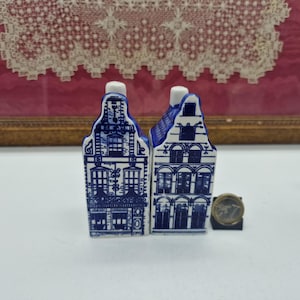 Può includere: Due casette in miniatura in ceramica con disegni blu e bianchi in stile Delft. Le case hanno facciate dettagliate, camini bianchi e sono accanto a una moneta. Lo sfondo presenta un merletto incorniciato.