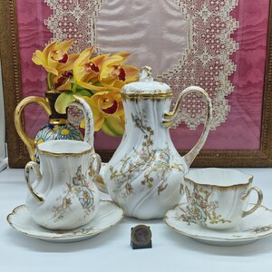 May include: A white porcelain tea set with gold detailing and floral patterns. The set comprises a teapot, two cups with saucers, and a creamer. A vase of yellow orchids is in the background. A coin is placed in front of the tea set.