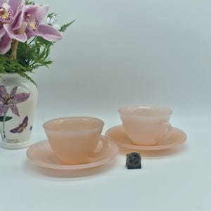 Könnte beinhalten: Zwei rosa Teetassen und Untertassen mit geripptem Design. Die Teetassen haben Henkel und stehen auf passenden Untertassen. Ein kleines, dunkles Objekt befindet sich zwischen den Untertassen. Eine Vase mit Blumen ist im Hintergrund.