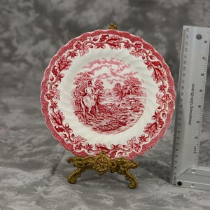 Può includere: Un piatto decorativo bianco con un design floreale e paesaggistico rosso, esposto su un supporto dorato. Il piatto ha un bordo smerlato e un'illustrazione dettagliata. Un righello è accanto al piatto.