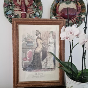 May include: Framed vintage print titled "La Moda" featuring two women in elegant Victorian-era dresses. The print is in a brown wooden frame and is displayed with decorative plates and a potted orchid.