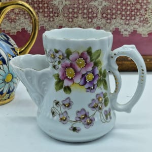 Puede incluir: Un tazón de afeitar de porcelana blanca con asa y jabonera incorporada. La taza está decorada con diseños florales rosas y morados pintados a mano y ribetes dorados. Un accesorio de baño vintage.