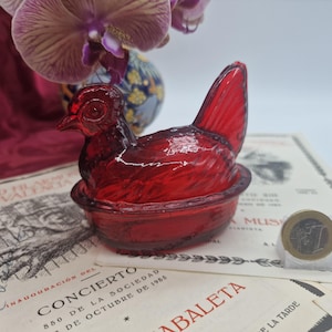 Puede incluir: Una gallina de cristal rojo rubí translúcido sobre un plato en forma de nido. La figura de la gallina presenta detalles de plumas moldeadas y una cola prominente. Una moneda de un euro está colocada junto al plato como escala de referencia.