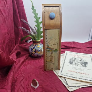 May include: A tall, rectangular wooden box with a light blue knob and a vertical panel featuring a printed illustration of a figure near a body of water and a mountain. The box is positioned next to a small round object and printed paper programs.