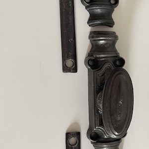 Könnte beinhalten: Ein Set aus dunkelgrauen Fensterbeschlägen aus Metall, bestehend aus zwei langen flachen Stangen mit kreisförmigen Löchern an den Enden, vier dekorativen Montagehalterungen mit Schraublöchern und einem zentralen Verschlussmechanismus.