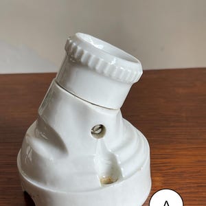 Puede incluir: Toma de corriente eléctrica de cerámica blanca con una toma de luz montada en el lateral. La toma tiene una superficie texturizada y un agujero para tornillos. La toma está inclinada y tiene un borde acanalado. La toma está etiquetada con "A".