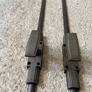 Könnte beinhalten: Zwei dunkelgraue Metalltürriegel mit rechteckigen Griffen und langen, zylindrischen Stangen. Jeder Riegel trägt die Buchstaben "LC" auf dem Griff. Die Riegel sind für einen sicheren Türverschluss konzipiert.