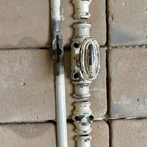 Puede incluir: Un mecanismo de cierre de ventana de metal blanco vintage con un asa ovalada central y una varilla vertical larga. Los herrajes presentan un acabado de pintura desgastada que revela metal oscuro, compuesto por varios segmentos.