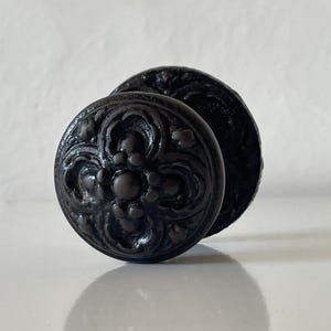 Può includere: Un pomello rotondo per mobili di colore marrone scuro con un motivo floreale in rilievo simmetrico sulla parte anteriore. Il pomello ha una finitura liscia e lucida e una piastra di base circolare, con dettagli stampati complessi.