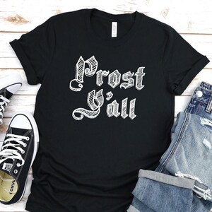 May include: Black t-shirt with the text "Prost Y'all" in white, stylized lettering. The shirt is paired with blue denim jeans and black canvas sneakers with white laces. A casual, everyday outfit.