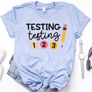 May include: Light blue t-shirt with the text "TESTING testing 1 2 3" and a yellow pencil graphic.