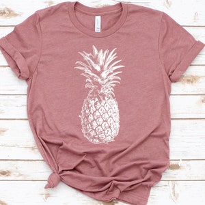 Pode incluir: Uma camiseta cor-de-rosa com um estampado de abacaxi branco.
