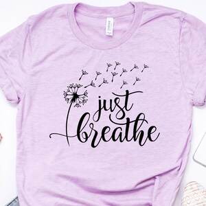Op de afbeelding: Een lichtpaars T-shirt met een zwarte paardenbloem afbeelding en de tekst "just breathe" in een cursief schrift.