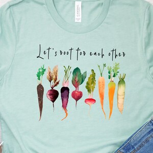 May include: A light blue t-shirt with the text "Let's root for each other" printed on the front. The shirt features a watercolor design of various vegetables, including carrots, beets, and radishes.