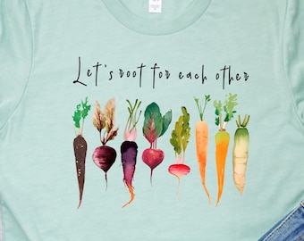 May include: A light blue t-shirt with the text "Let's root for each other" printed on the front. The shirt features a watercolor design of various vegetables, including carrots, beets, and radishes.