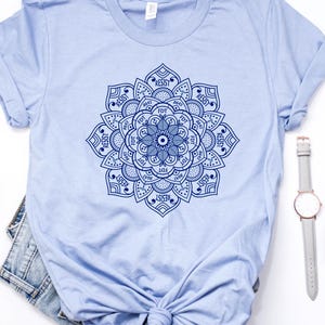 May include: A light blue t-shirt featuring a detailed blue mandala design. The shirt is styled with a pair of white flats, denim shorts, a wristwatch, and sunglasses. The shirt's design includes the word "RESIST".