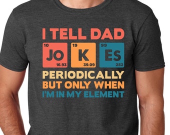 Op de afbeelding: Donkergrijs t-shirt met de tekst "I TELL DAD JOKES PERIODICALLY BUT ONLY WHEN I'M IN MY ELEMENT" in oranje, blauw en geel. Het woord "JOKES" wordt weergegeven als elementen uit het periodiek systeem.
