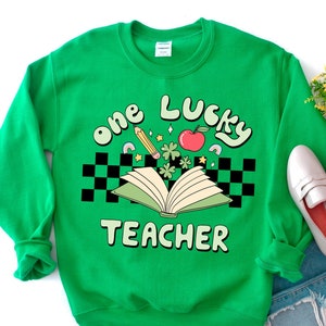May include: Green sweatshirt with the text "one lucky teacher" in a retro font. The design includes a book, a pencil, a red apple, and four-leaf clovers.