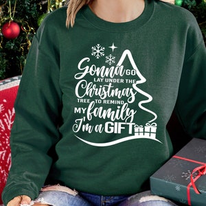 May include: A green sweatshirt with a white Christmas tree design. The text on the tree reads "Gonna go lay under the Christmas tree to remind my family I'm a gift".