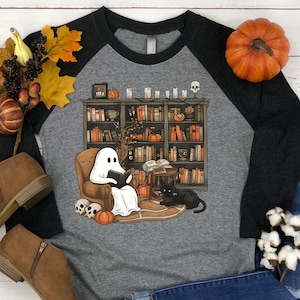 May include: A gray and black baseball tee shirt with a ghost reading a book in a chair in front of a bookshelf. The ghost is white and has a black cat sitting next to it. The bookshelf is filled with books and has a few pumpkins on the shelves.