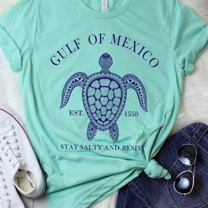 Puede incluir: Una camiseta verde claro con un dise&ntilde;o de tortuga azul marino y el texto "GULF OF MEXICO EST. 1550 STAY SALTY AND RESIST." Zapatillas blancas y pantalones cortos de mezclilla est&aacute;n al lado de la camisa.