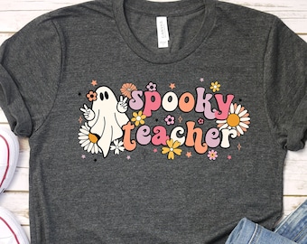 Op de afbeelding: Een donkergrijs T-shirt met een spook en de tekst "spooky teacher" in kleurrijke retro letters omringd door bloemen.