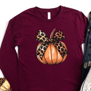 May include: A maroon long-sleeved t-shirt with a graphic of a pumpkin with a leopard print bow.