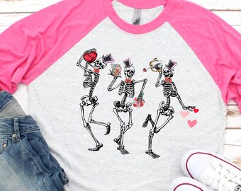 May include: A pink and grey long-sleeved baseball tee shirt with a graphic of three skeletons dancing and holding hearts. The skeletons are wearing top hats and have floral accents.