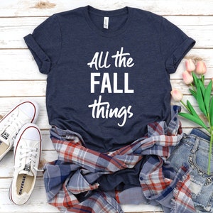 May include: A navy blue t-shirt with the text "All the FALL things" in white lettering. The shirt is laid out on a white wooden surface with a plaid shirt, denim shorts, and pink tulips.