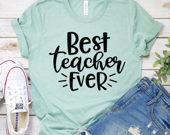 Op de afbeelding: Een lichtblauw T-shirt met de tekst "Best teacher ever" in zwarte letters. Het shirt ligt plat op een houten oppervlak met een paar witte sneakers en een spijkerbroek.