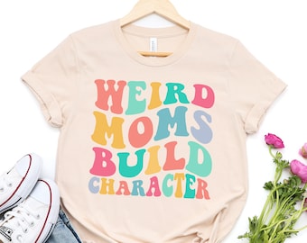 May include: A beige t-shirt with the text "Weird Moms Build Character" in rainbow colors. The text is in a retro font style.