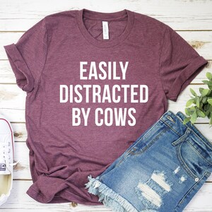 May include: A maroon t-shirt with the text "EASILY DISTRACTED BY COWS" in white lettering. The shirt is laid out on a white wooden surface with a pair of white sneakers and denim shorts.