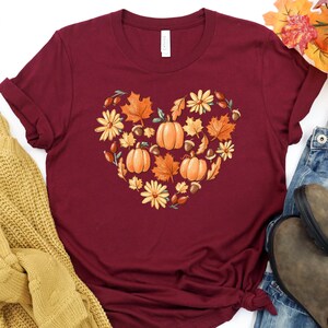 May include: A maroon t-shirt with a heart-shaped design made of fall leaves, pumpkins, acorns, and flowers.