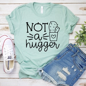 May include: A light green t-shirt with the text "NOT a hugger" and a black line drawing of a cactus in a pot. The shirt is laying on a white wooden surface with a pair of white canvas shoes and a pair of blue denim shorts.