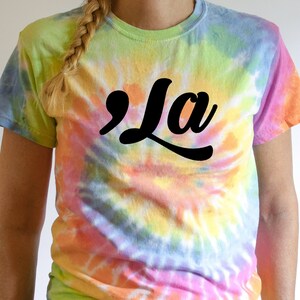 May include: A tie-dye t-shirt with a black and white graphic of the word "'La" in a retro font.