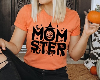 May include: A woman wearing a black hat with gold stars and a bright orange t-shirt with the text "MOMSTER" in black. The shirt has a witch hat, moon, stars, spider, and pumpkin graphic. She is holding a small orange pumpkin.