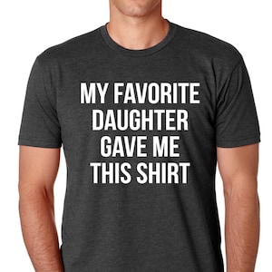 K&ouml;nnte beinhalten: Dunkelgraues T-Shirt mit dem wei&szlig;en Text "MY FAVORITE DAUGHTER GAVE ME THIS SHIRT". Das Shirt hat einen Rundhalsausschnitt und scheint aus einem weichen, bequemen Material zu sein.