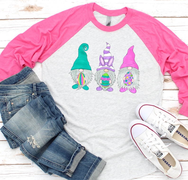 May include: A pink and grey long-sleeved baseball tee shirt with a design of three gnomes holding Easter eggs. The gnomes are wearing green, purple, and pink hats.