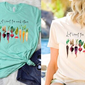 May include: Two light-colored t-shirts with the text "Let's root for each other" and a watercolor illustration of various vegetables, including carrots, radishes, and beets.