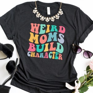 Pode incluir: Uma camiseta cinza escura com o texto "Weird Moms Build Character" em cores do arco-&iacute;ris. O texto est&aacute; em um estilo de fonte retr&ocirc;.