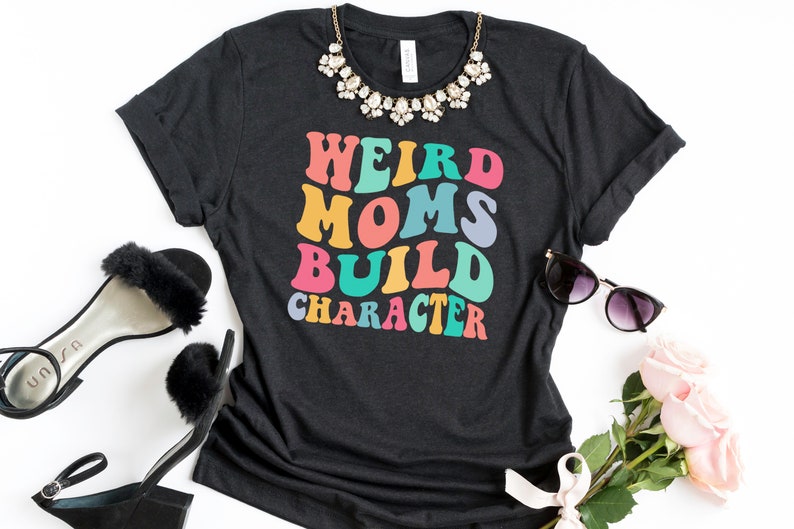 Pode incluir: Uma camiseta cinza escura com o texto "Weird Moms Build Character" em cores do arco-&iacute;ris. O texto est&aacute; em um estilo de fonte retr&ocirc;.