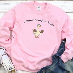 May include: Pink sweatshirt with the text "outnumbered by boys." A cartoon goose wearing a hat and holding a football and a toy truck is printed on the front. White sneakers and blue jeans are also in the image.