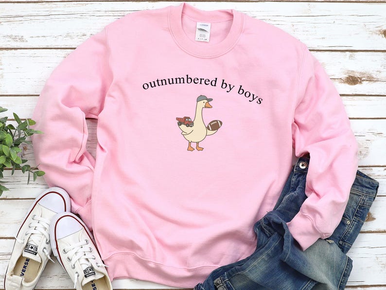 May include: Pink sweatshirt with the text "outnumbered by boys." A cartoon goose wearing a hat and holding a football and a toy truck is printed on the front. White sneakers and blue jeans are also in the image.