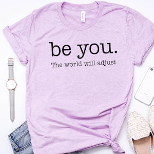 Puede incluir: Una camiseta lila con el texto negro "be you. The world will adjust". La imagen incluye un reloj gris, un smartphone blanco, zapatos color crema y unos vaqueros azules.