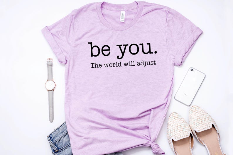 Puede incluir: Una camiseta lila con el texto negro "be you. The world will adjust". La imagen incluye un reloj gris, un smartphone blanco, zapatos color crema y unos vaqueros azules.