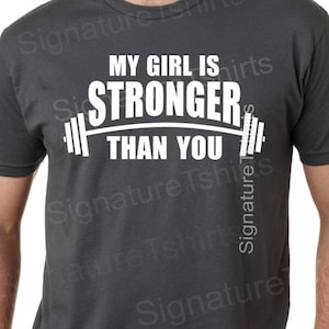 May include: A dark gray t-shirt with white text that reads "My girl is stronger than you" with a barbell graphic.